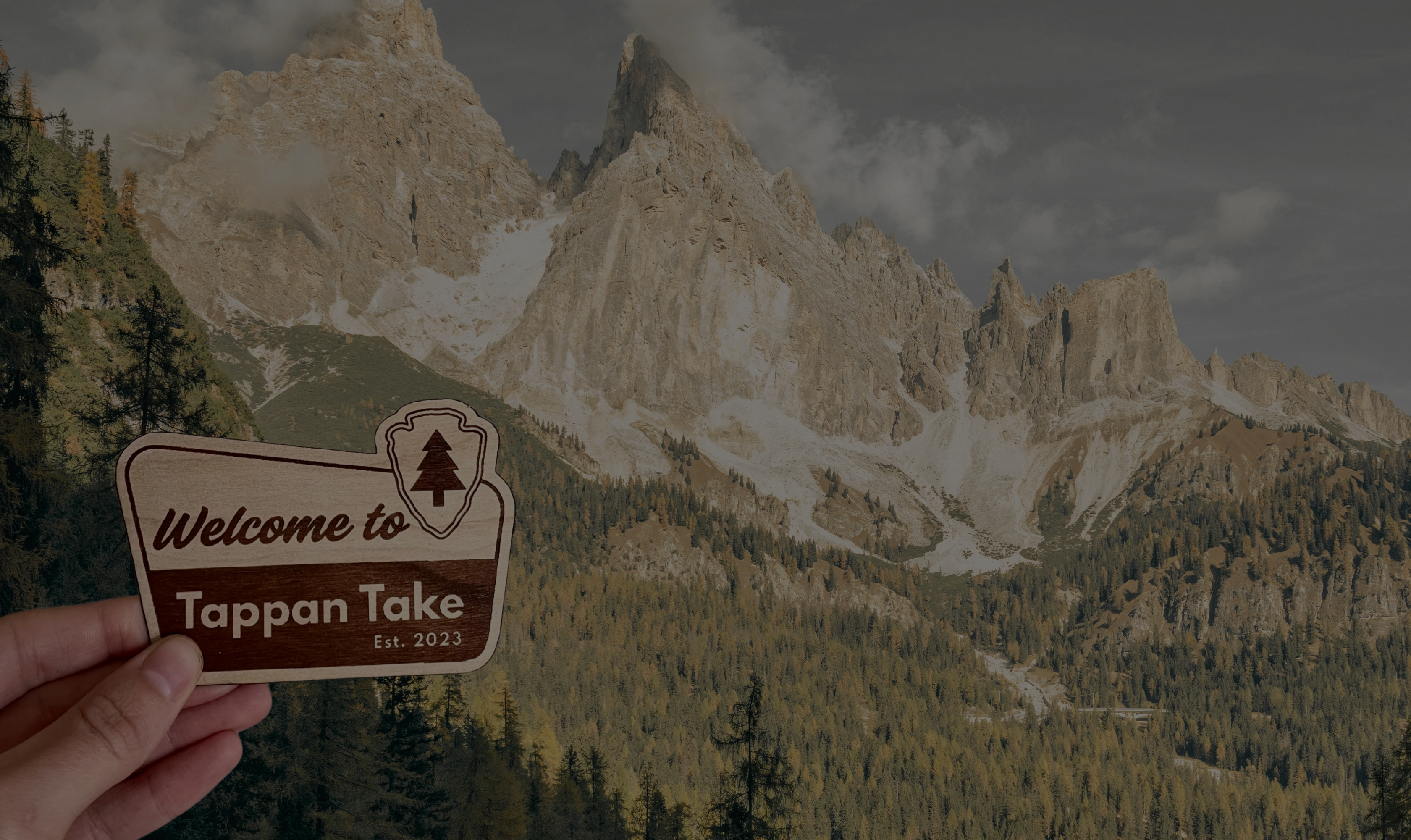Hand holding a Tappan Take 'Welcome to Tappan Take' wood sticker in front of dramatic mountain peaks
